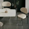 Modern dining room with a white marble table, beige chairs with black legs, and a bottle with two wine glasses on the table, plus a tall plant in the background.