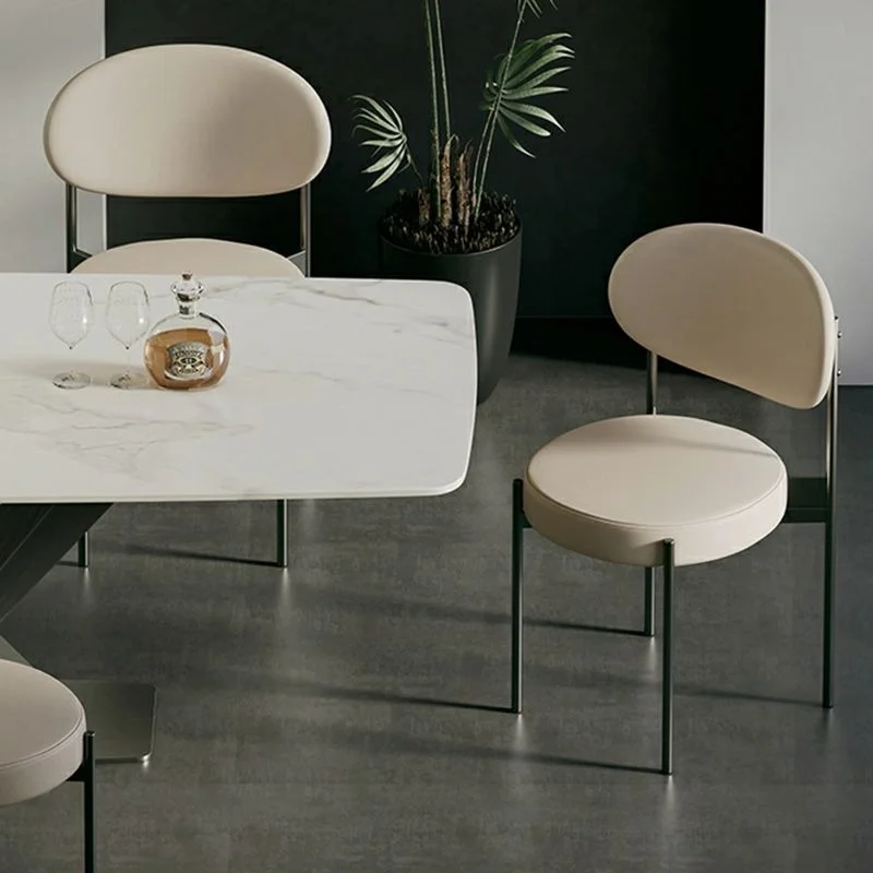 Modern dining room with a white marble table, beige chairs with black legs, and a bottle with two wine glasses on the table, plus a tall plant in the background.