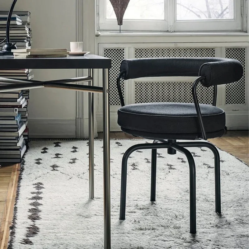 A black padded office chair at a glass-topped desk in a bright home office with a stack of books nearby and a patterned rug.