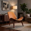 Mid-century orange leather lounge chair with curved wood arms on a beige rug, beside a floor lamp in a cozy living room.