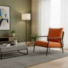 Orange leather armchair with black metal frame sits beside a gray coffee table on a textured rug in a modern living room with a green wall, floor lamp, abstract wall art, and potted plants.