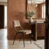 Beige upholstered chair with black legs beside a dark wood console, vase of eucalyptus, and a stack of books under a large gold-framed mirror on terracotta walls.