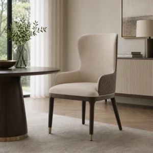 Beige upholstered dining chair with a quilted side panel and dark wood legs with brass tips beside a round table in a bright living room.