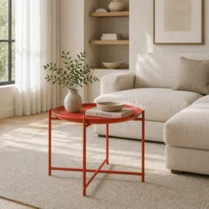 Red round metal side table in a sunlit living room holds a vase with greenery, a bowl, and a book beside a beige sofa.