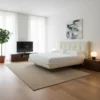 Bright minimalist bedroom with a cream upholstered bed, two brown nightstands, round lamps, abstract art above the headboard, and a TV on a low console.