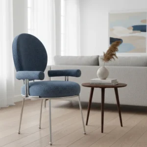 Blue textured upholstered chair with metal legs sits beside a round wooden side table holding a small vase with pampas grass in a bright, minimalist living room.