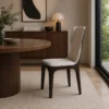 Round wooden dining table with a beige upholstered chair in a modern, sunlit dining room.