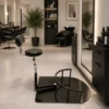 Modern hair salon interior with black styling chairs, large mirrors, and potted plants; foreground shows a circular-base stool-like chair accessory.