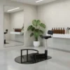 Minimalist salon station with a black hydraulic chair on a circular platform, mirror, and white shelf with brown bottles.
