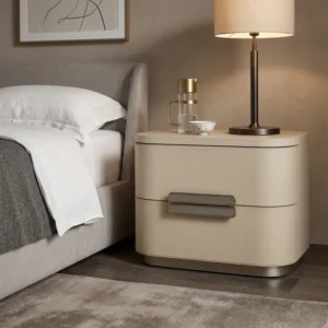 Beige curved two-drawer nightstand beside a bed with a lamp, carafe, and teacup on top in a neutral bedroom setting.