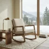Wooden rocking chair with beige cushions beside a rustic end table and brass lamp, facing a snowy mountain view.