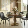 Round wooden dining table with a brass pedestal base, six black upholstered chairs, and green dishware set with a floral centerpiece.