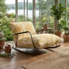 Cozy sunroom with a mustard-yellow upholstered rocking chair, surrounded by plants, near large windows overlooking a lake.