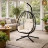 Black metal hanging egg chair with cream cushions in a bright living room with plants and a beige sofa nearby.