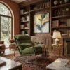 Cozy library corner with a green lounge chair, built-in wooden shelves, a framed leaf painting, and a lit table lamp.