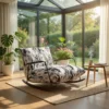 Rocking chair with a black-and-white pattern on a beige rug in a sunlit conservatory; a small table holds a steaming mug and a book beside lush plants outside the glass walls.