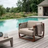 Poolside seating on a wooden deck with a beige cushioned chair and gray blanket beside a bright pool, plus a table with a glass and sunglasses.