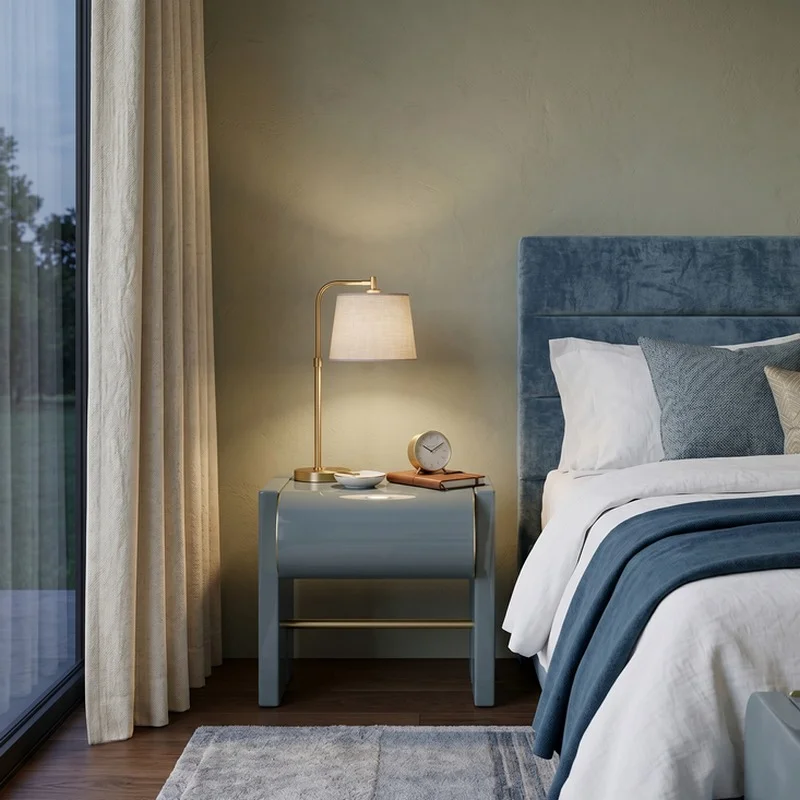 Bedside table with a brass lamp, clock, and books beside a blue upholstered headboard in a softly lit bedroom.