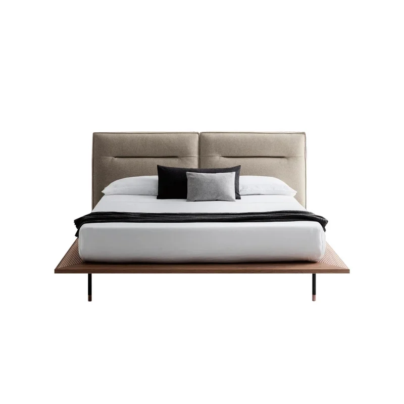 Modern platform bed with beige upholstered headboard, white bedding, and two accent pillows (black and gray). The low wooden frame and slim legs are visible beneath the mattress.