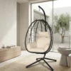 Black egg-shaped hanging chair with cream cushions on a stand in a bright modern living room by a large window.