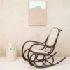 Modern black rocking chair with a woven seat in a light studio, beside a round side table holding a pitcher and books, with an abstract framed painting on the wall.