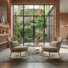 Modern living room with two beige rocking armchairs facing a glass coffee table on a patterned rug, large grid windows and greenery outside.