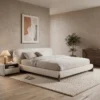 6-96458 Modern beige bedroom with a low upholstered bed, neutral bedding, nightstand and lamp, on a woven rug with a potted plant nearby.