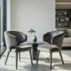 Two gray upholstered lounge chairs face a small round marble table with a vase and leaf in a bright, modern living room.