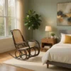 6-96671 Sunlit bedroom with a wooden rocking chair by a large window, a bed with white bedding and a mustard pillow, and a nightstand with a lamp and plant nearby.