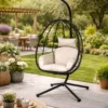 Outdoor hanging egg chair with beige cushions on a black stand in a garden patio setting.