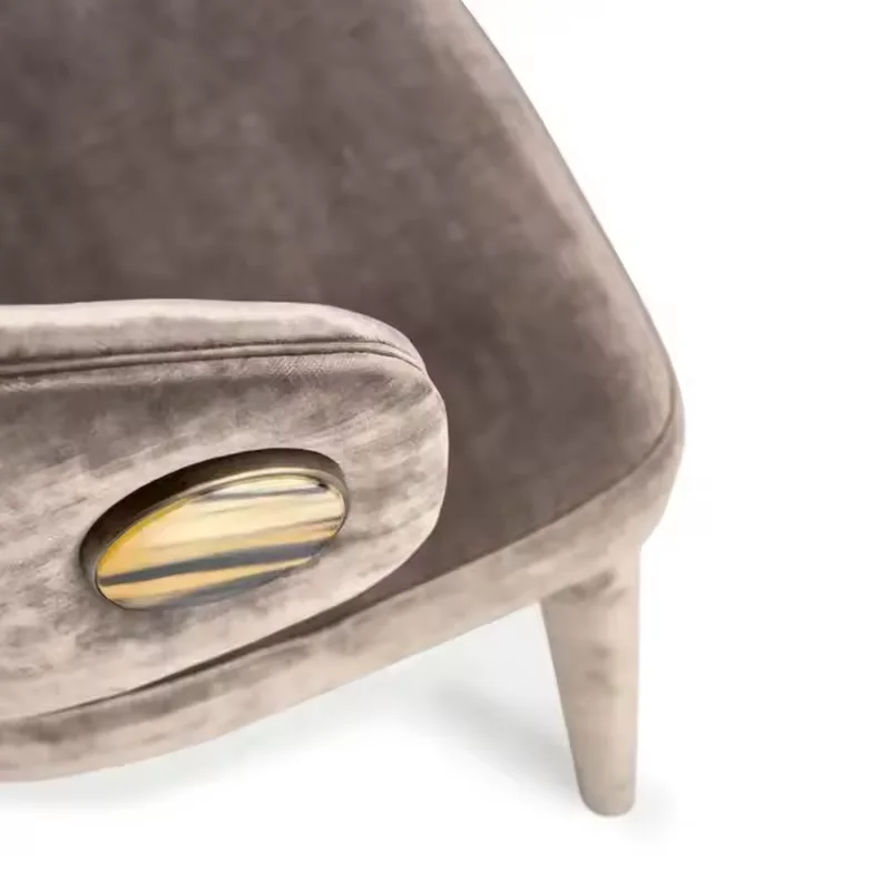 Close-up of a taupe velvet upholstered chair featuring a curved arm and a gold oval button detail.