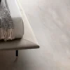 Beige sofa edge with a fringed knit blanket draped over the arm and a dark leg visible beneath the seat