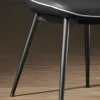 Close-up of three metal chair legs supporting a black cushioned seat on a wooden floor, with a white edge detail visible at the top edge of the seat.