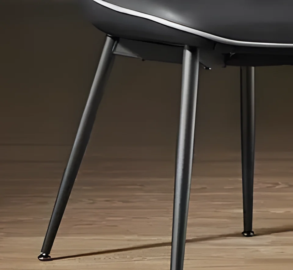 Close-up of three metal chair legs supporting a black cushioned seat on a wooden floor, with a white edge detail visible at the top edge of the seat.