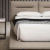 Bed with beige upholstered headboard, white linens, and a textured throw blanket in a modern bedroom setup with a glass-topped nightstand nearby