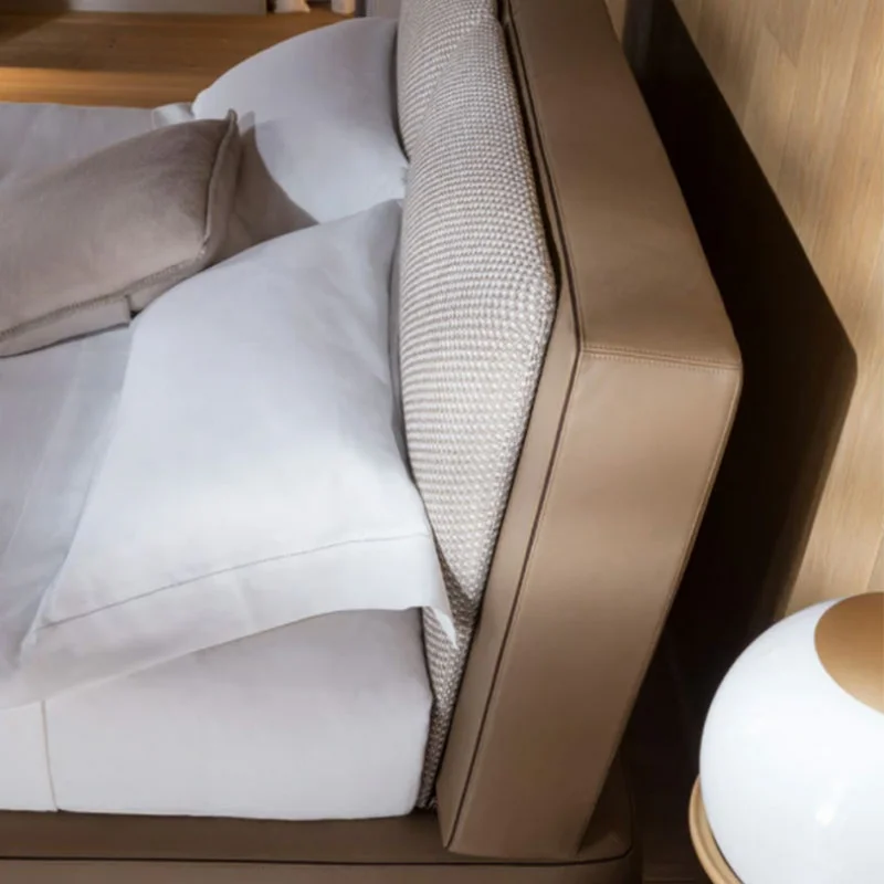 Close-up of a bed with white bedding, beige upholstered headboard, and a beige throw on a light wood floor. A rounded bedside lamp is partially visible on the right.