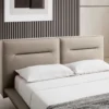 Beige upholstered bed with two white pillows and white sheets, against a striped vertical-panel wall with a framed abstract art piece above.