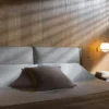 Modern bedroom with wood paneling, an upholstered headboard, white linens, a brown throw pillow, and a lit bedside lamp on the right.