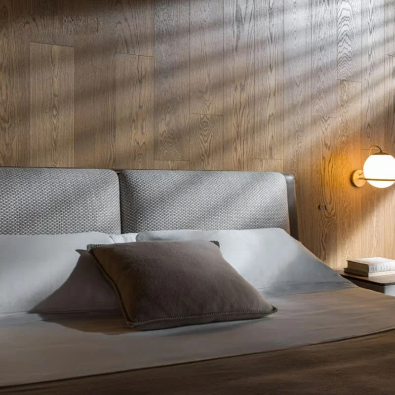Modern bedroom with wood paneling, an upholstered headboard, white linens, a brown throw pillow, and a lit bedside lamp on the right.
